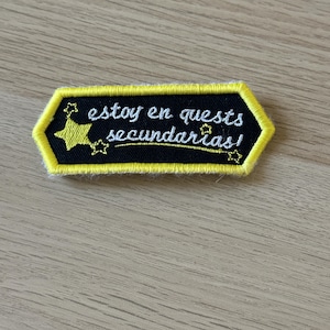 Puede incluir: Un parche bordado negro y amarillo con el texto "estoy en quests secundarias!". El parche tiene un borde amarillo y detalles de estrellas. El texto está en escritura blanca.