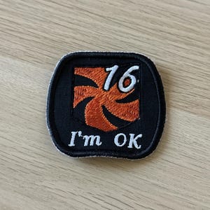 Puede incluir: Un parche negro con borde blanco. El parche presenta un diseño naranja estilizado y el número "16" en blanco. Debajo del diseño, el texto "I'm OK" está bordado en blanco.