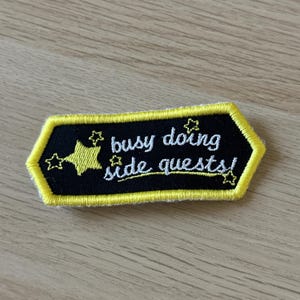 Puede incluir: Parche bordado con fondo negro y borde amarillo. El parche presenta el texto "busy doing side quests!" en escritura blanca, con detalles de estrellas amarillas.