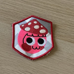 Puede incluir: Parche bordado con un personaje de dibujos animados de hongo con una cara rosa, mejillas rojas y una gorra de hongo roja con lunares blancos. El parche tiene un borde rojo y un fondo a cuadros gris y blanco.