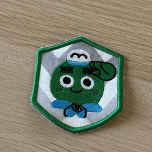 Puede incluir: Parche bordado con un personaje de dibujos animados verde con uniforme de marinero, saludando. El personaje tiene grandes ojos negros y una cara sonriente. El parche está bordeado en verde y sobre un patrón de chevron gris y blanco.