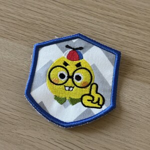 Puede incluir: Parche bordado con un personaje de dibujos animados amarillo con gafas, un gorro rojo y azul y un dedo levantado. El parche tiene un borde azul y un fondo de chevron blanco y gris.