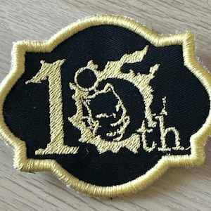 Puede incluir: Parche bordado negro con ribete y letras doradas. El parche dice "15th" en una fuente estilizada. El parche tiene un borde decorativo.