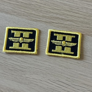 Puede incluir: Dos parches bordados cuadrados, con borde dorado sobre fondo negro. El diseño dorado presenta la letra "H" con una calavera alada en el centro. Los parches están sobre una superficie de madera clara.