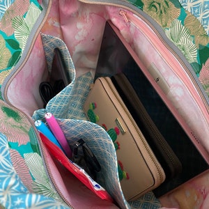 May include: Open tote bag with a floral pattern in teal, pink, and white. The interior features multiple pockets holding a tablet, wallet, pens, and other items. The bag has a gray zipper and a pink interior lining.