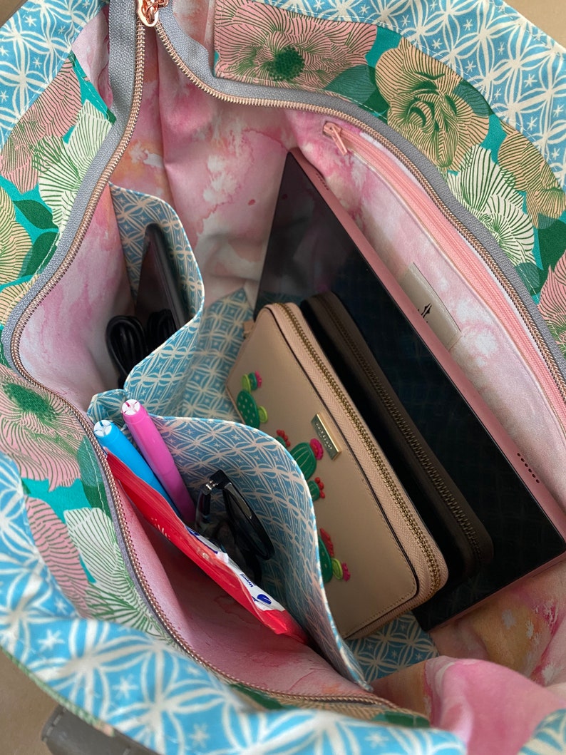 May include: Open tote bag with a floral pattern in teal, pink, and white. The interior features multiple pockets holding a tablet, wallet, pens, and other items. The bag has a gray zipper and a pink interior lining.
