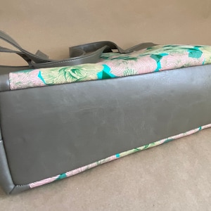 May include: A gray tote bag with a rectangular shape and two handles. The top section features a colorful floral pattern with pink, green, and blue accents. The bag is made of a smooth, leather-like material.
