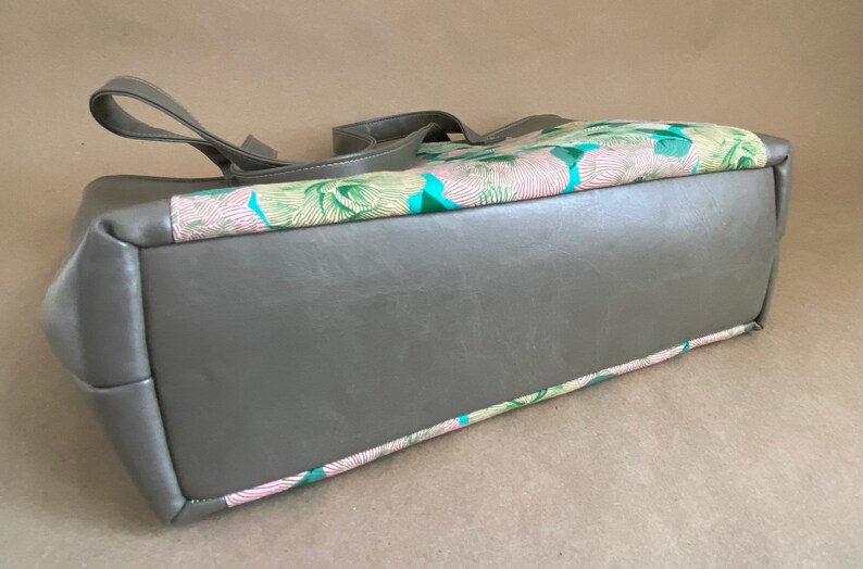 May include: A gray tote bag with a rectangular shape and two handles. The top section features a colorful floral pattern with pink, green, and blue accents. The bag is made of a smooth, leather-like material.