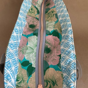 May include: Open tote bag with a floral interior. The interior features a teal background with pink and white flowers. The bag has a gray zipper with a rose gold pull. The exterior is a dark gray.