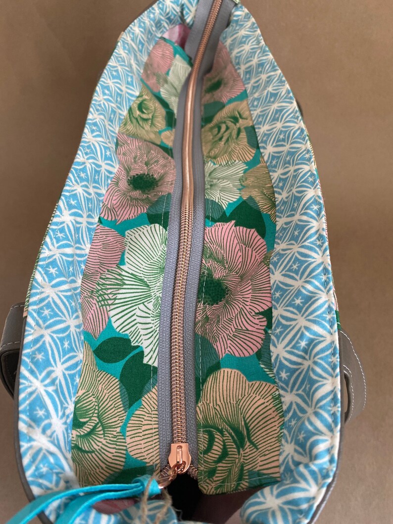 May include: Open tote bag with a floral interior. The interior features a teal background with pink and white flowers. The bag has a gray zipper with a rose gold pull. The exterior is a dark gray.