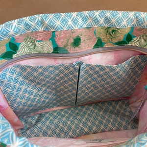 May include: Open view of a floral tote bag interior. The bag features a light blue geometric pattern on the trim and pockets, with a floral print in shades of pink, green, and teal. The interior has multiple pockets and a zipper closure.