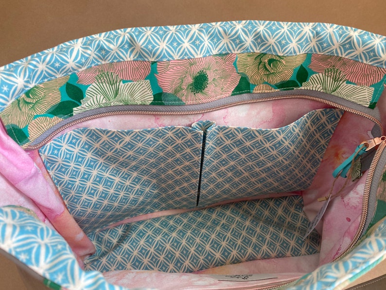 May include: Open view of a floral tote bag interior. The bag features a light blue geometric pattern on the trim and pockets, with a floral print in shades of pink, green, and teal. The interior has multiple pockets and a zipper closure.