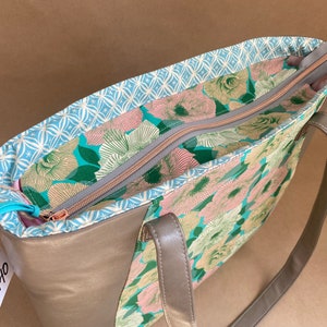 May include: A beige tote bag with a floral pattern interior and a light blue patterned lining. The bag has a zipper closure and gray handles. The interior features a teal, pink, and green floral design.