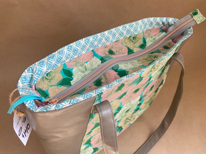 May include: A beige tote bag with a floral pattern interior and a light blue patterned lining. The bag has a zipper closure and gray handles. The interior features a teal, pink, and green floral design.