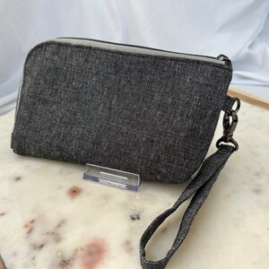 May include: A gray zippered pouch with a black strap and a silver clasp. The pouch is made of a textured fabric.