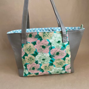May include: A gray tote bag with a floral pattern. The bag features a central panel with pink, green, and white flowers on a teal background. Gray handles and sides complete the design. The interior lining has a blue and white geometric pattern.