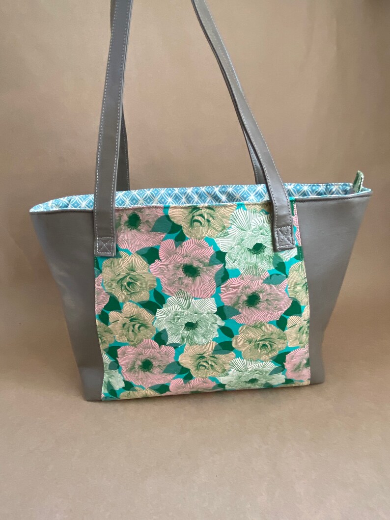 May include: A gray tote bag with a floral pattern. The bag features a central panel with pink, green, and white flowers on a teal background. Gray handles and sides complete the design. The interior lining has a blue and white geometric pattern.