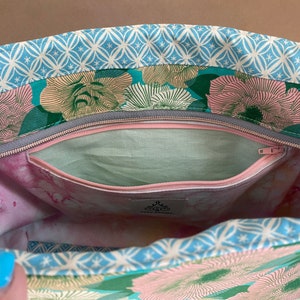 May include: Open view of a floral tote bag interior. The bag features a light blue geometric patterned trim, with a teal and pink floral design. A zippered pocket is visible, along with a light green interior lining and a pink and white marbled pattern.