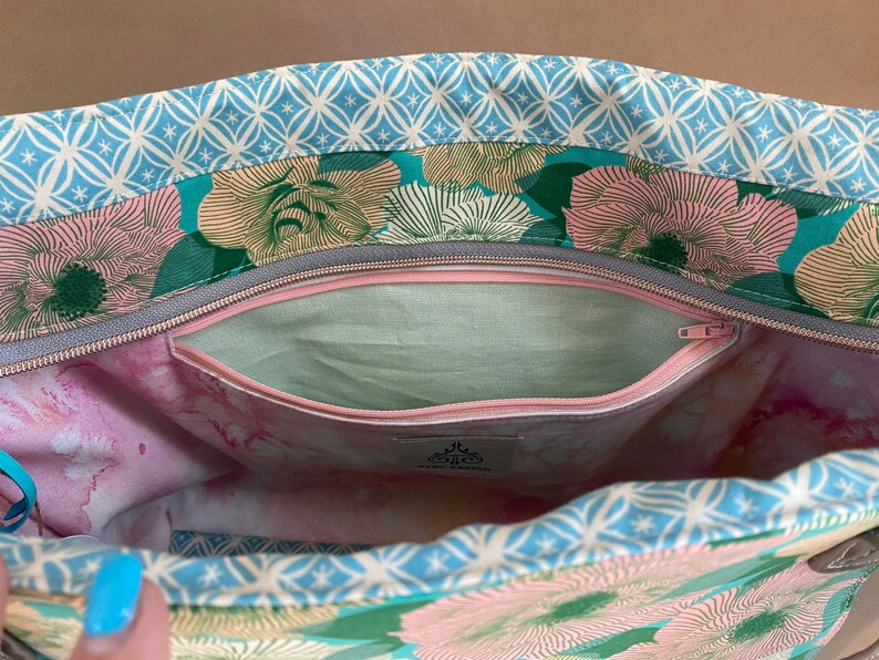 May include: Open view of a floral tote bag interior. The bag features a light blue geometric patterned trim, with a teal and pink floral design. A zippered pocket is visible, along with a light green interior lining and a pink and white marbled pattern.
