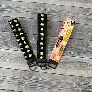 May include: Three keychains with different patterns. The first keychain has a black background with yellow skulls and flowers. The second keychain has a black background with yellow flowers and skulls. The third keychain has a pink and yellow background with a geometric pattern.