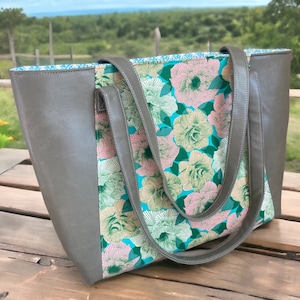 May include: A gray tote bag with a floral pattern. The bag features a gray exterior with a floral print panel in shades of pink, green, and teal. The bag has matching gray handles and a teal interior lining.