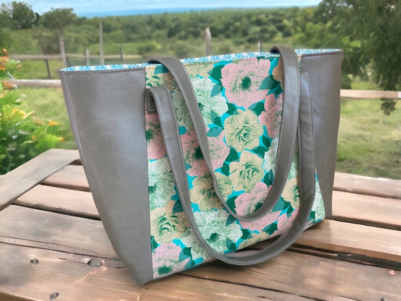 May include: A gray tote bag with a floral pattern. The bag features a gray exterior with a floral print panel in shades of pink, green, and teal. The bag has matching gray handles and a teal interior lining.