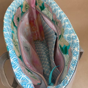 May include: Open view of a silver tote bag with a floral patterned lining. The interior features multiple pockets and a zippered closure. The lining has a blue and white geometric pattern, with pink and green floral accents.