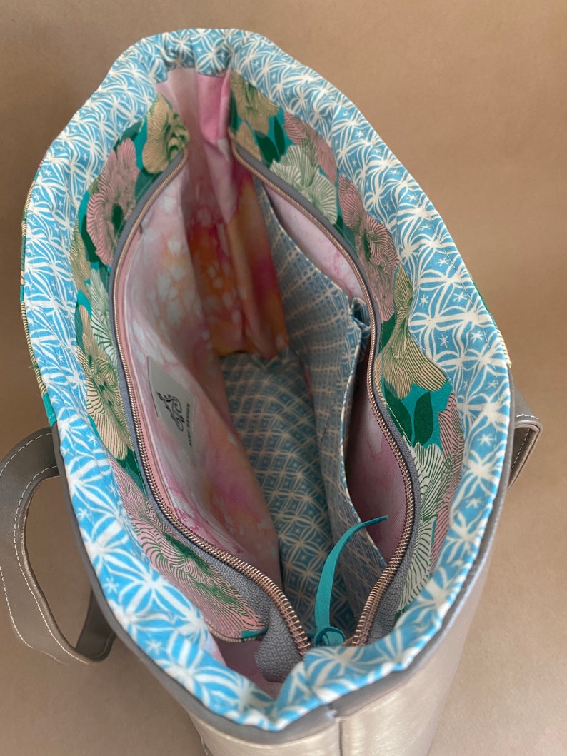 May include: Open view of a silver tote bag with a floral patterned lining. The interior features multiple pockets and a zippered closure. The lining has a blue and white geometric pattern, with pink and green floral accents.