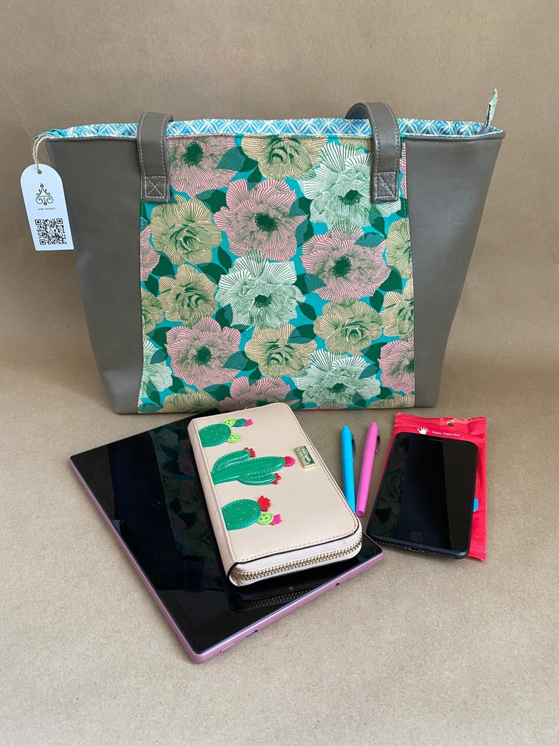 May include: A gray tote bag with a floral pattern, a pink tablet, a beige wallet with cactus designs, a smartphone, and two pens. The tote bag has gray handles and sides, and a floral center panel with pink, green, and white flowers on a teal background.