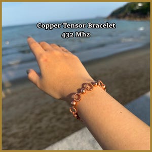 Op de afbeelding: Een koperen tensorarmband met afwisselend gladde bolvormige kralen en gedraaide cirkelvormige schakels. De tekst Copper Tensor Bracelet 432 Mhz staat boven de armband, die om een pols wordt gedragen tegen een wazige achtergrond van de kust.