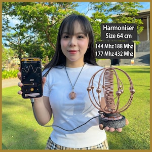 May include: A copper-colored Harmoniser sculpture with a spherical wire frame and central coil, mounted on a dark speckled base. The device is connected by a black cable to a smartphone displaying a frequency generator app with the text 432 Hz.