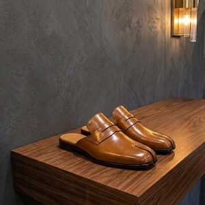 Op de afbeelding: Een paar lichtbruine leren instappers met een opvallend ontwerp met gespleten neus, een gladde gepolijste afwerking en een klassieke penny loafer-band over de wreef. Deze hielvrije schoenen hebben een lage hak en een gestroomlijnd model.