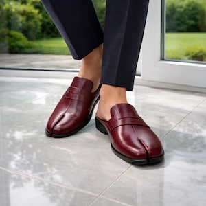 May include: Burgundy leather slip-on shoes with a split-toe design. The shoes have a low heel and a strap across the top. The shoes are worn with navy blue pants. The shoes are on a gray tiled floor.