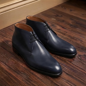 Könnte beinhalten: Ein Paar marineblaue Chukka-Stiefel aus Leder mit glatter Oberfläche, abgerundeter Zehenpartie und einer Schnürung mit zwei Ösen sowie dünnen schwarzen Schnürsenkeln. Die Stiefel haben ein beiges Innenfutter und eine dunkle Sohle mit klarer Form.