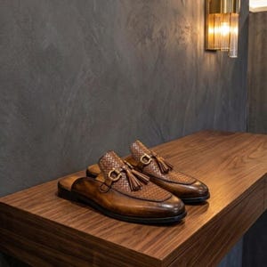 Peut inclure: Une paire de mocassins ouverts à l'arrière en cuir marron avec une texture tressée sur le dessus, un pompon décoratif et un mors en métal doré. Les chaussures présentent une finition patinée et un petit talon, dans un style à enfiler.