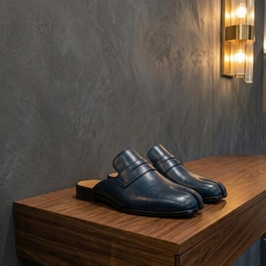 Könnte beinhalten: Ein Paar dunkelblaue Leder-Mules mit einem klassischen Penny-Steg über dem Vorderblatt, einer abgerundeten Zehenpartie und einem niedrigen Blockabsatz. Die Slipper haben eine glatte Oberfläche und eine strukturierte Form.