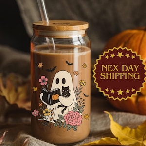 May include: A glass beverage cup with a bamboo lid and straw, featuring a Halloween-themed illustration of a ghost reading a book with a black cat. The cup is filled with a brown liquid and surrounded by pumpkins and fall leaves.