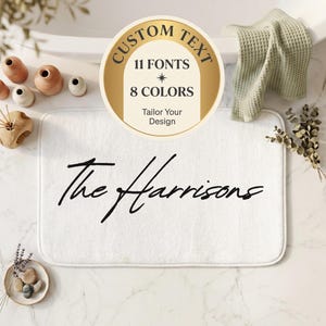 May include: A rectangular white bath mat featuring the black script text The Harrisons centered on the surface. A circular graphic overlay displays the text CUSTOM TEXT 11 FONTS 8 COLORS Tailor Your Design in a gold and white arch layout.