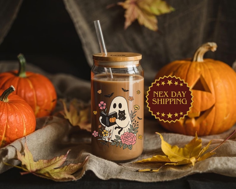 May include: A glass beverage cup with a bamboo lid and straw, featuring a Halloween-themed illustration of a ghost reading a book with a black cat. The cup is filled with a brown liquid and surrounded by pumpkins and fall leaves.