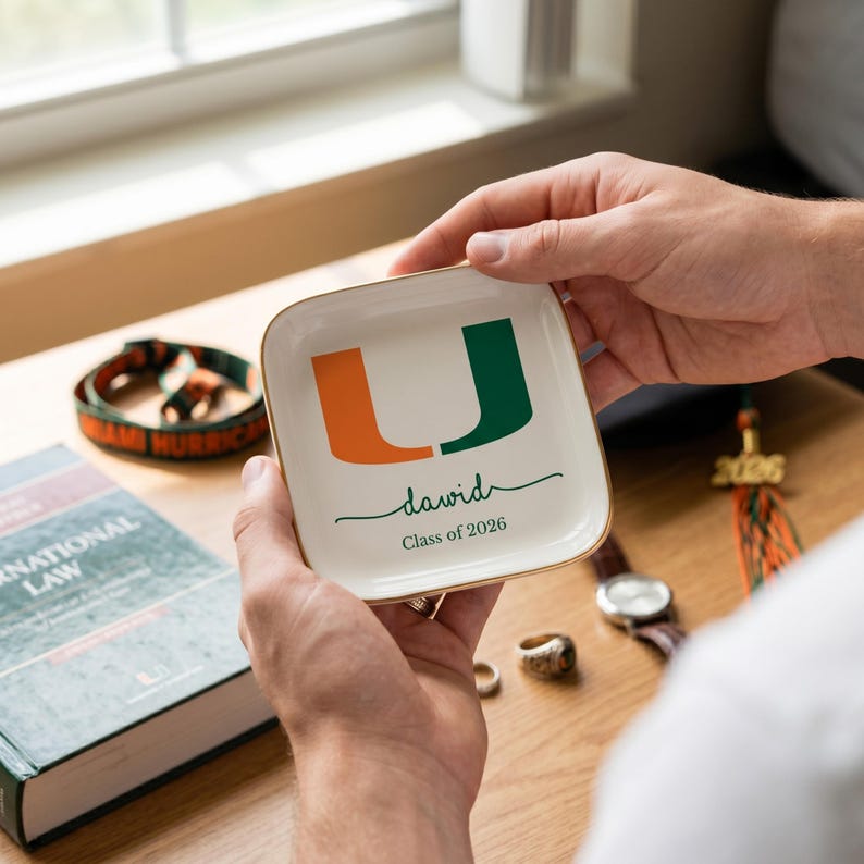 Puede incluir: Un platillo cuadrado blanco con borde dorado, con el logotipo "U" de la Universidad de Miami en naranja y verde. El platillo est&aacute; personalizado con el nombre "David" y "Class of 2026" en escritura verde.