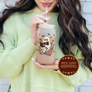 May include: A frosted glass can-shaped tumbler with a bamboo lid and straw. The tumbler features a Halloween-themed design with a ghost reading a book, a black cat, flowers, and bats. The tumbler is held by a person wearing a light green sweater.