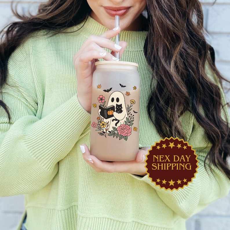 May include: A frosted glass can-shaped tumbler with a bamboo lid and straw. The tumbler features a Halloween-themed design with a ghost reading a book, a black cat, flowers, and bats. The tumbler is held by a person wearing a light green sweater.