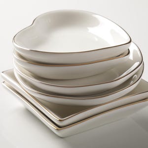 Puede incluir: Una pila de platos de cer&aacute;mica blanca con borde dorado. La pila incluye platos en forma de coraz&oacute;n y cuadrados. Los platos est&aacute;n apilados, creando una disposici&oacute;n visualmente atractiva. Los platos est&aacute;n limpios y tienen un acabado brillante.