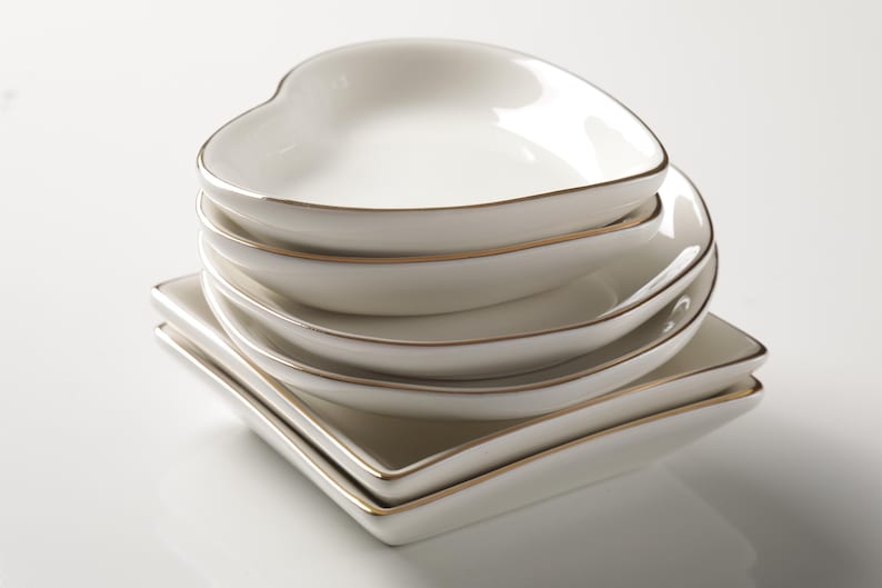 Puede incluir: Una pila de platos de cer&aacute;mica blanca con borde dorado. La pila incluye platos en forma de coraz&oacute;n y cuadrados. Los platos est&aacute;n apilados, creando una disposici&oacute;n visualmente atractiva. Los platos est&aacute;n limpios y tienen un acabado brillante.