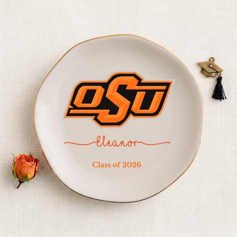Puede incluir: Un plato de cer&aacute;mica blanco con borde dorado, con las letras "OSU" en naranja y negro. El plato est&aacute; personalizado con el nombre "Eleanor" y "Class of 2026". Un peque&ntilde;o dije de gorro de graduaci&oacute;n y una rosa naranja est&aacute;n al lado.