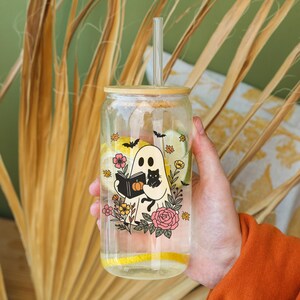 May include: A clear glass tumbler with a bamboo lid and straw, featuring a Halloween-themed design. The design includes a ghost reading a book, a black cat, flowers, and bats. The tumbler is filled with water and lemon slices.