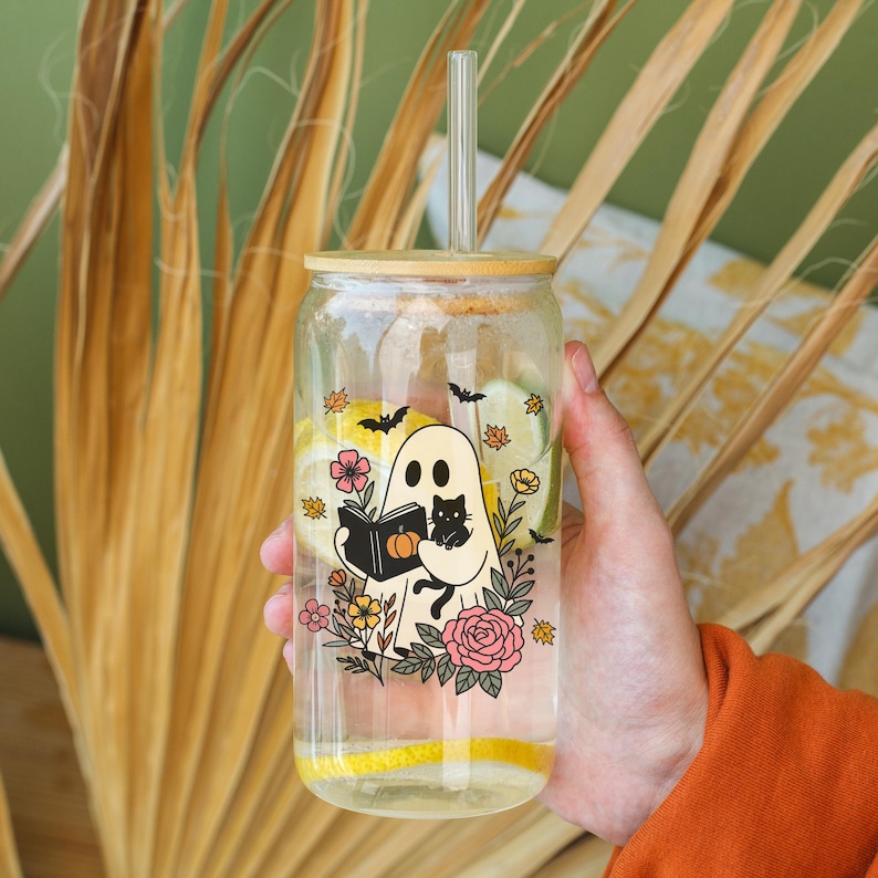 May include: A clear glass tumbler with a bamboo lid and straw, featuring a Halloween-themed design. The design includes a ghost reading a book, a black cat, flowers, and bats. The tumbler is filled with water and lemon slices.