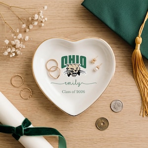 Puede incluir: Un joyero de cer&aacute;mica blanca en forma de coraz&oacute;n con borde dorado, con el logotipo de la Universidad de Ohio y el nombre "emily" con "Class of 2026". El plato contiene anillos y pendientes. Un birrete y un diploma est&aacute;n en el fondo.