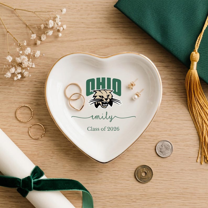 Puede incluir: Un joyero de cer&aacute;mica blanca en forma de coraz&oacute;n con borde dorado, con el logotipo de la Universidad de Ohio y el nombre "emily" con "Class of 2026". El plato contiene anillos y pendientes. Un birrete y un diploma est&aacute;n en el fondo.
