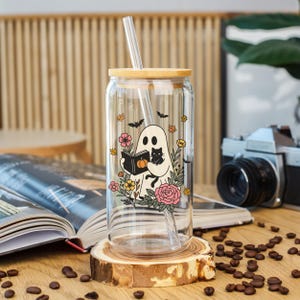 May include: A clear glass can-shaped tumbler with a bamboo lid and straw. The tumbler features a whimsical ghost design holding a book and a black cat, surrounded by flowers and bats. The tumbler is displayed on a wooden coaster.